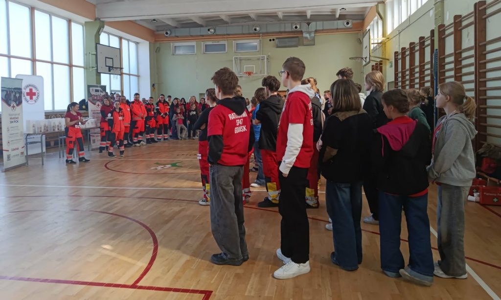 W sali gimnastycznej rzędy uczniów biorących udział w mistrzostwach PCK z PP. W tle banery PCK, organizatorzy i ratownicy.