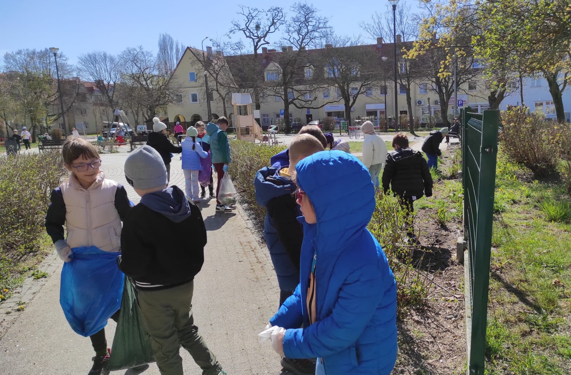 Grupa dzieci porządkuje plac zabaw. Część z nich trzyma worki, a inne wrzucają do nich śmieci. W tle budynki mieszkalne i wysokie drzewa.
