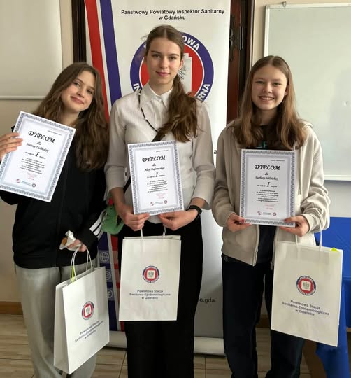 3 uczennice stoją obok siebie i uśmiechają się, trzymając dyplomy. Każda z nich ma w ręku torbę z nagrodami. W tle widoczny jest baner Powiatowej Stacji Sanitarno-Epidemiologicznej w Gdańsku.