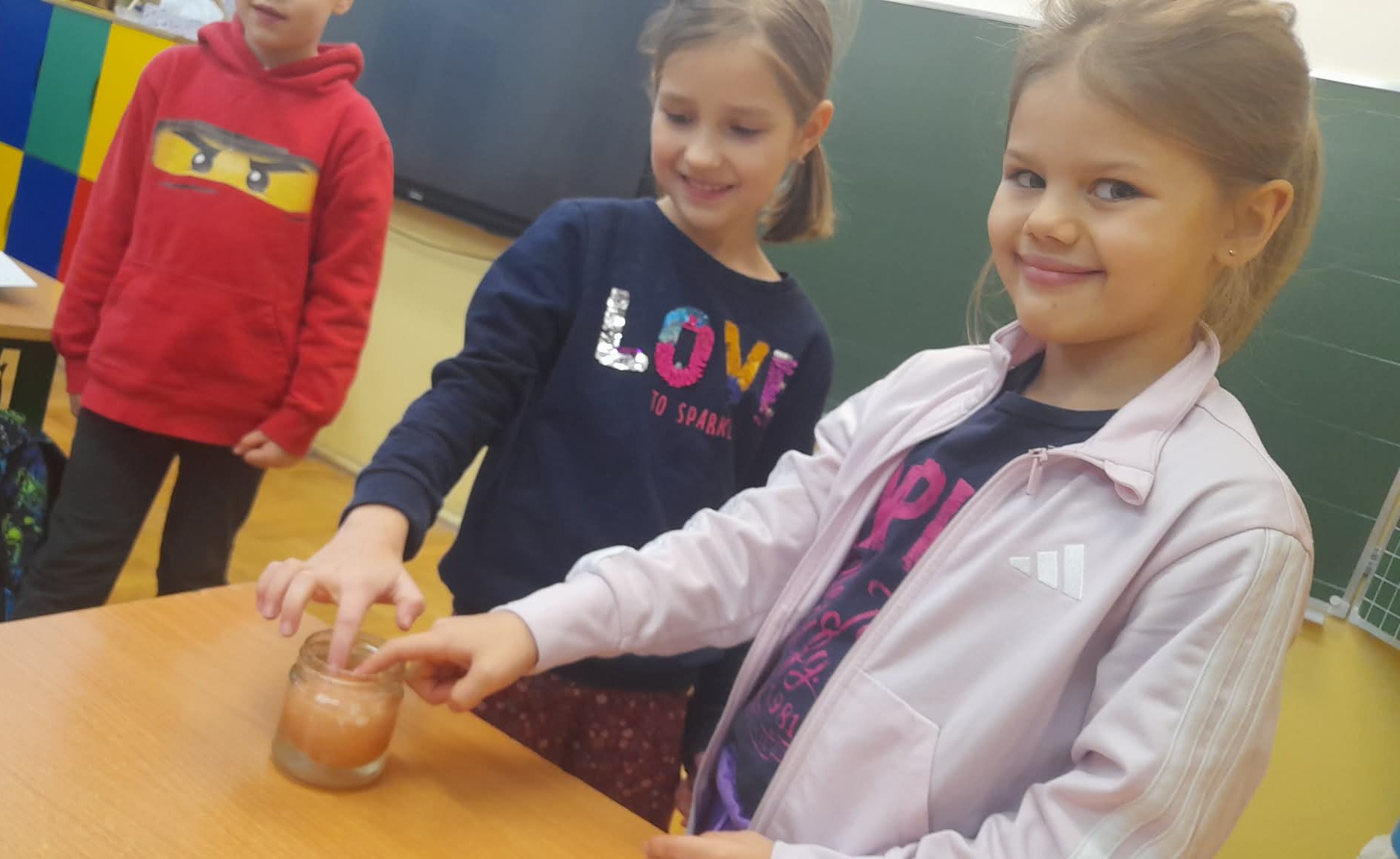 W sali lekcyjnej dwie dziewczynki wkładające place do słoika z jajkiem stojącego na ławce, w tle chłopiec.
