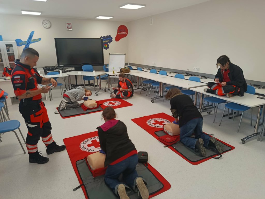 W sali na podłodze leżą trzy fantomy. Przed nimi uczennice robiące RKO. Obok ratownicy oceniając ich działania.