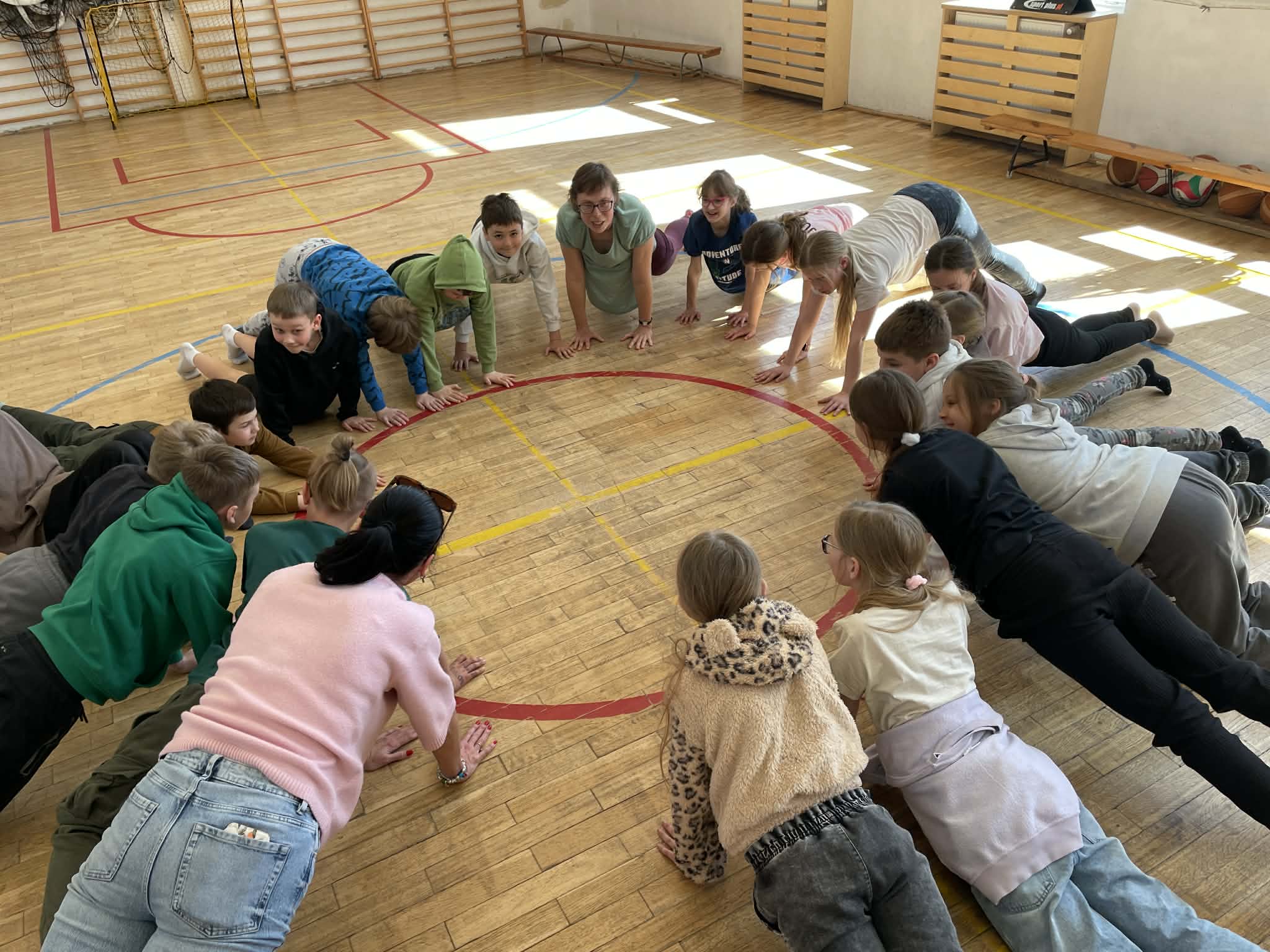 Uczniowie wraz z instruktorką tworzą duży krąg na środku sali gimnastycznej, wykonując ćwiczenie w podporze przodem (plank/pozycja deski) na drewnianym parkiecie. Uczestnicy są zwróceni twarzami do wnętrza koła, wyznaczonego przez czerwone linie boiska. Przez duże okna wpada jasne światło słoneczne.