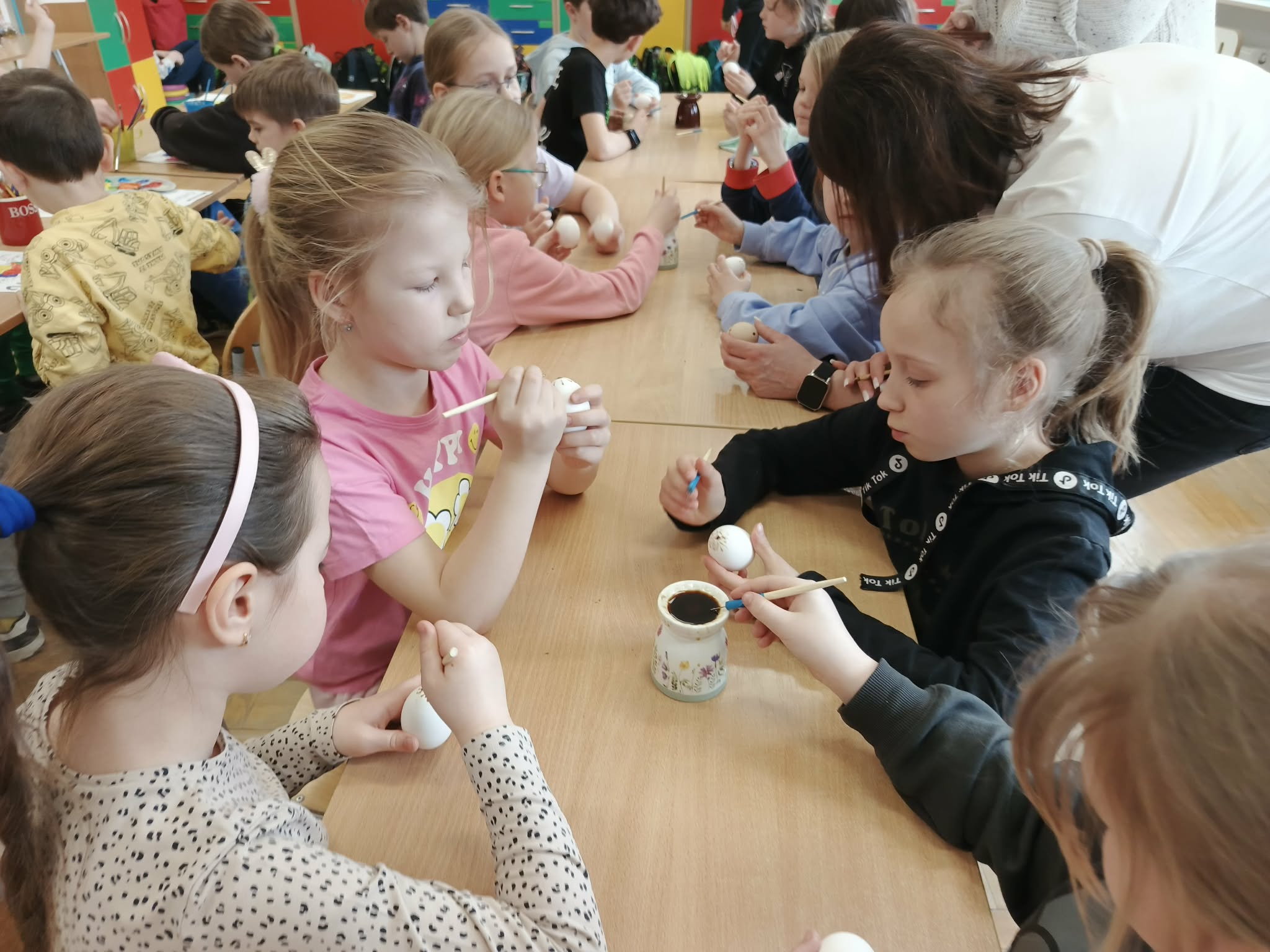 Dzieci siedzące przy stołach podczas zdobienia pisanek. Każde dziecko trzyma białe jajko i ma patyczek, którym nanoszą wzory z wosku.