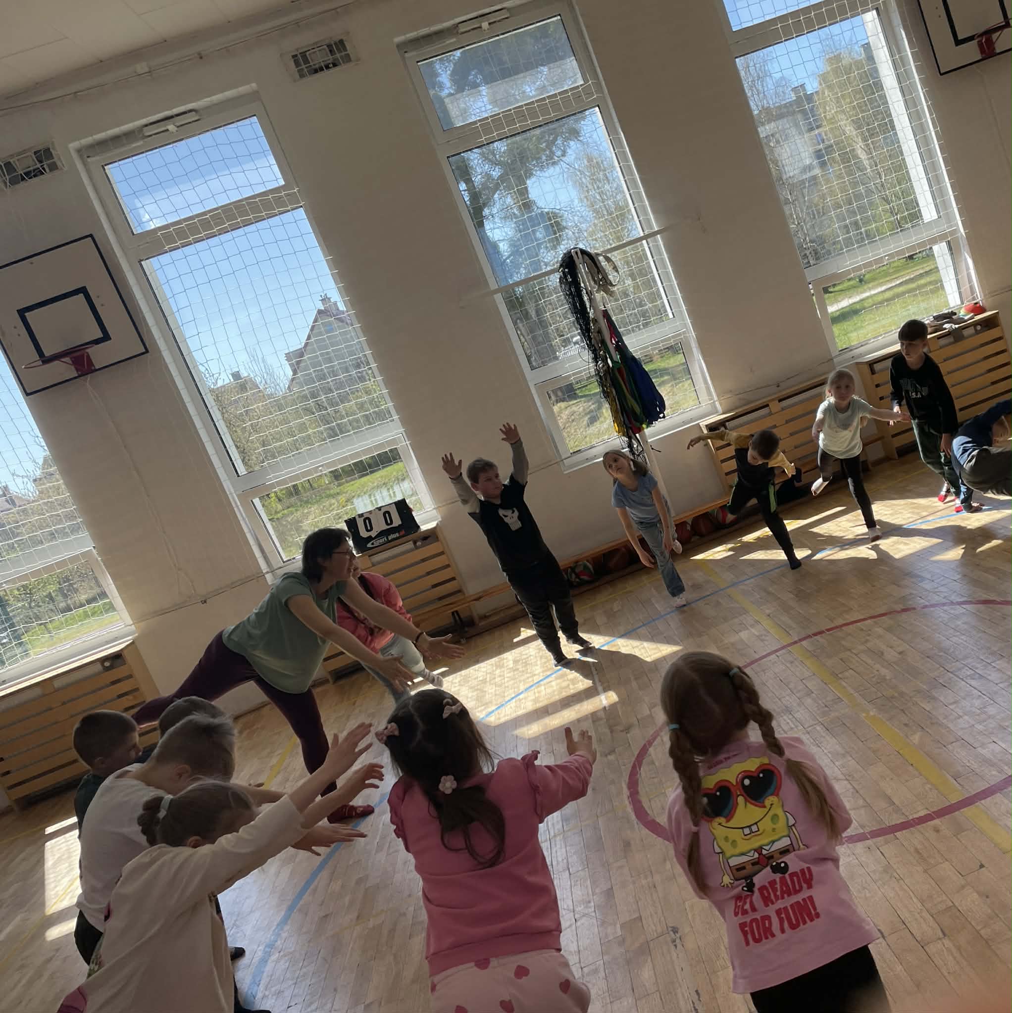 Grupa dzieci w sali gimnastycznej wykonuje stojące ćwiczenie ruchowe, unosząc ręce i wyciągając je przed siebie. Prowadząca, w turkusowej koszulce, stoi po lewej stronie, instruując grupę. Na pierwszym planie widać dziewczynkę w różowej bluzie z nadrukiem SpongeBoba. Promienie słońca oświetlają parkiet.