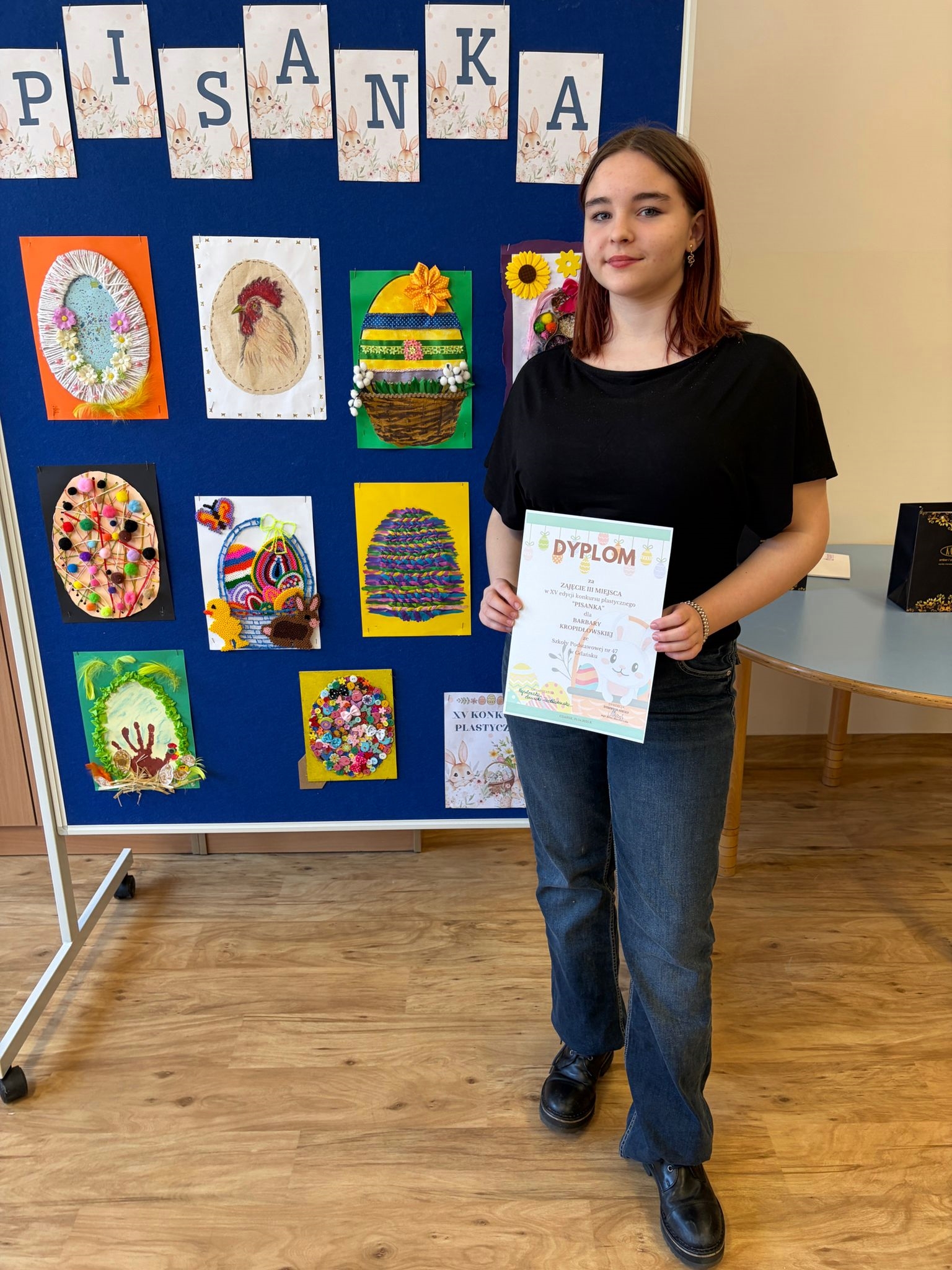 Portret młodej uśmiechniętej dziewczyny w czarnym t-shircie i jeansach, stojącej na tle niebieskiej tablicy z napisem "PISANKA". Dziewczyna trzyma w dłoniach dyplom za zajęcie III miejsca w konkursie plastycznym. Na tablicy obok niej wyeksponowane są kolorowe, wielkanocne prace plastyczne przedstawiające pisanki wykonane różnymi technikami.