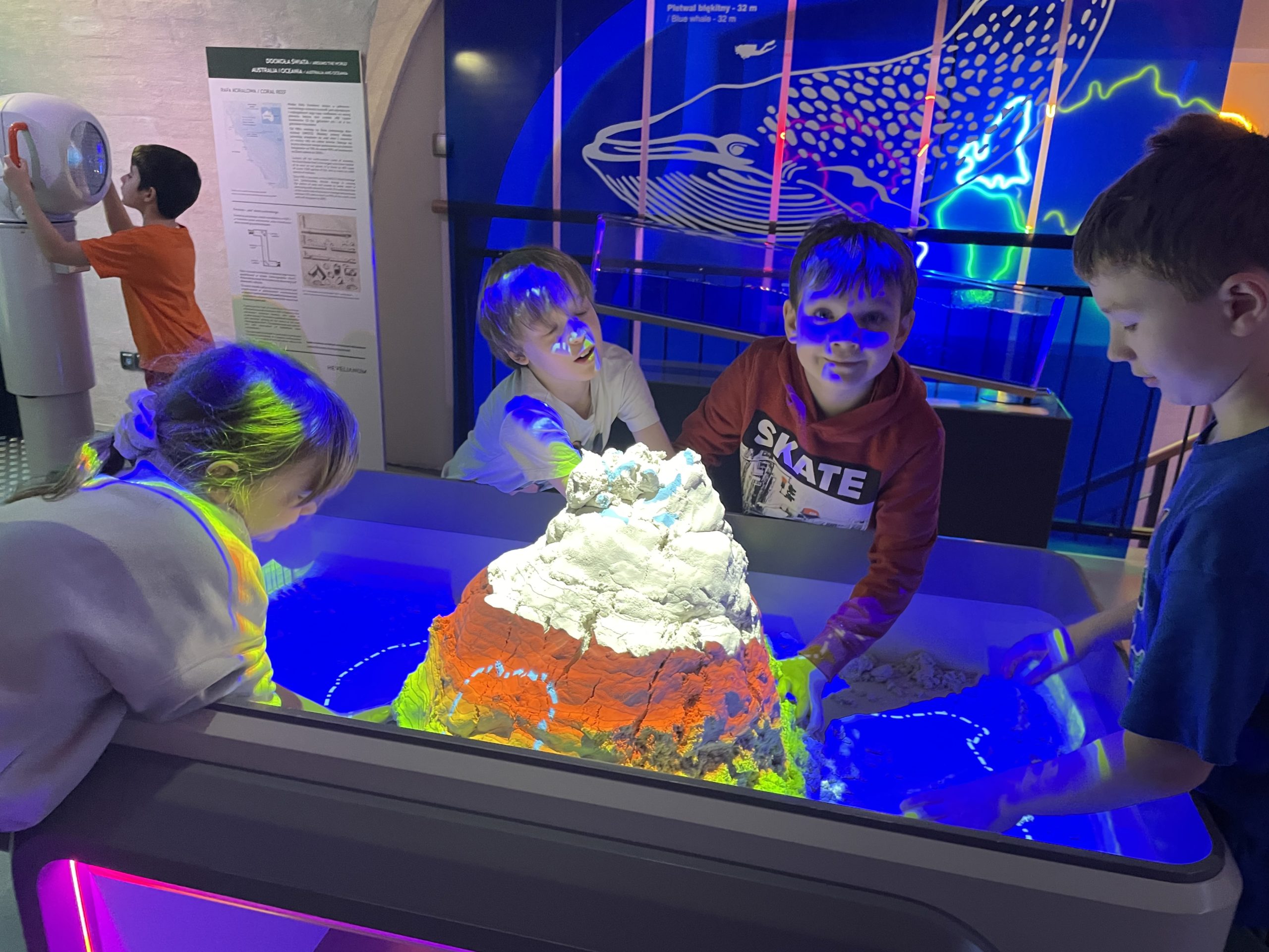 Czwórka dzieci stoi przy stanowisku z piaskiem o buduje górę. W tle jest chłopiec patrzący przez lornetkę.