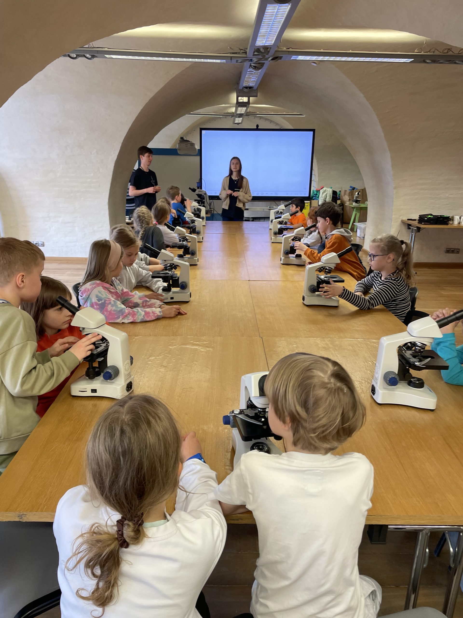 20 dzieci siedzi przy stole , przed nimi stoją mikroskopy. Na drugi planie stoi 2 prowadzących warsztaty.