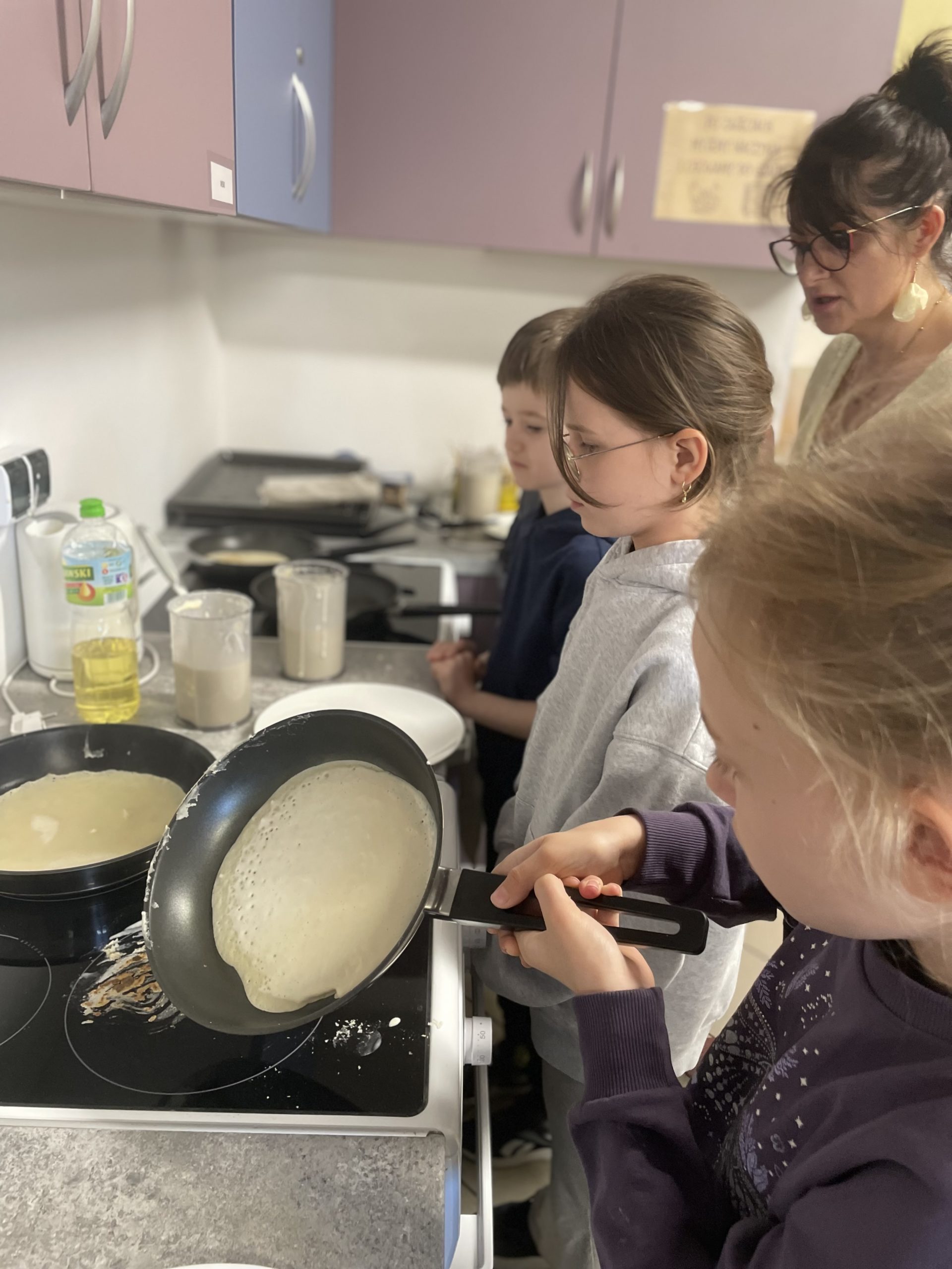 Troje dzieci stoi przy kuchenkach w rękach mają patelnie. Z tyłu stoi nauczycielka.