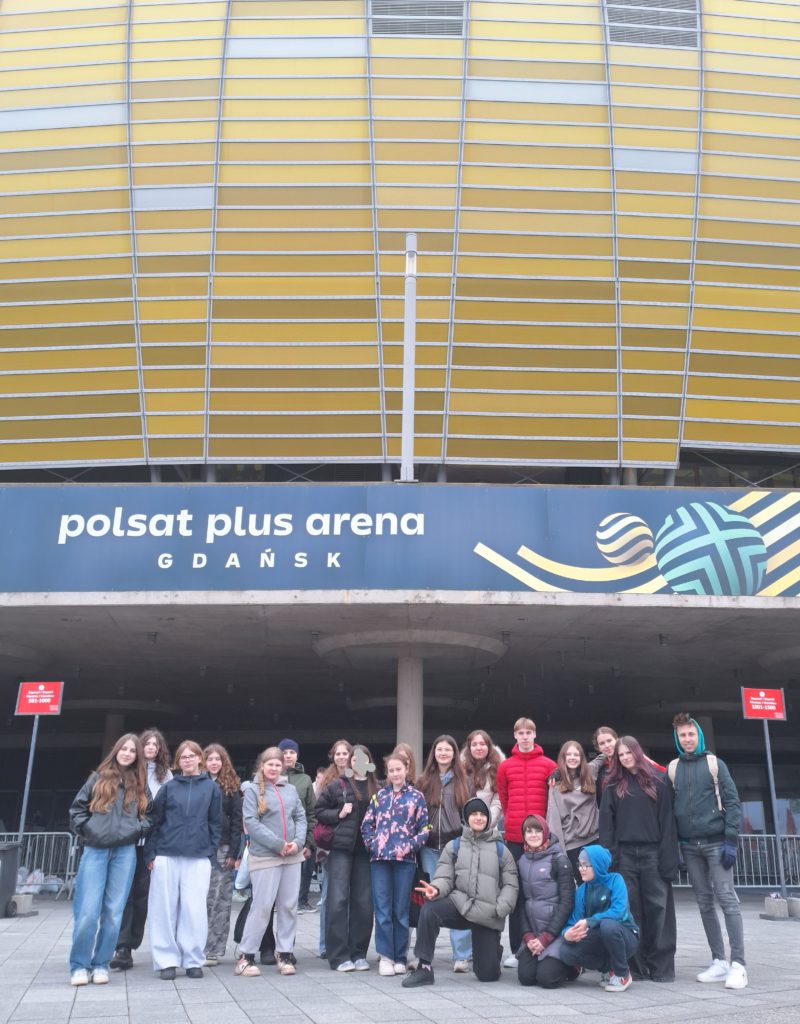 Grupa uczniów – wolontariuszy stoi przed wejściem do hali Polsat Plus Arena w Gdańsku. Ubrani w kurtki i bluzy, pozują wspólnie do zdjęcia na tle charakterystycznej elewacji budynku.