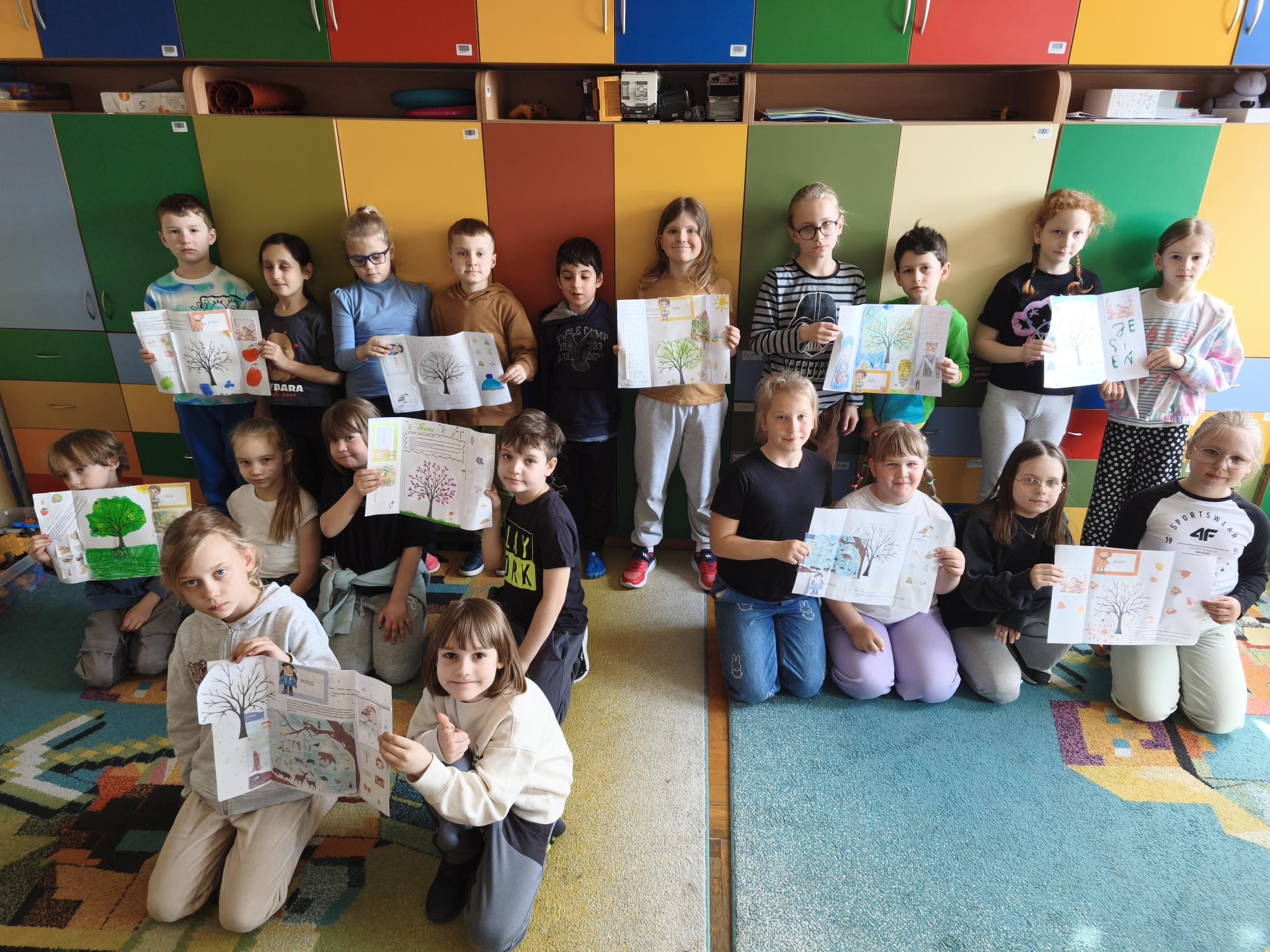 Na zdjęciu widać grupę dzieci w sali szkolnej, ustawionych w dwóch rzędach. Trzymają swoje prace plastyczne z rysunkami drzew i przyrody, patrząc w stronę aparatu. W tle są kolorowe szafki, a na podłodze leży wzorzysty, barwny dywan.