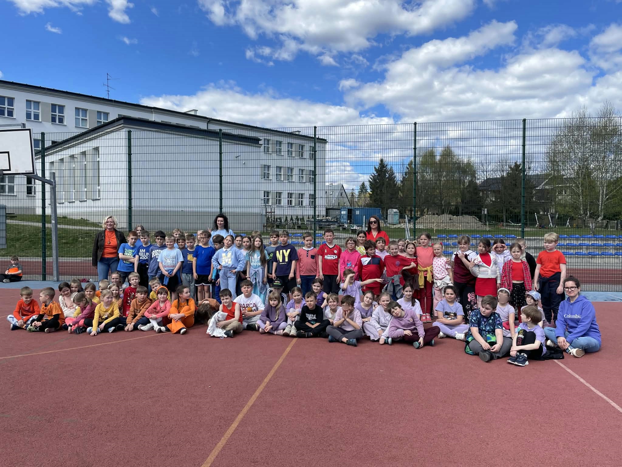 Duża grupa dzieci w różnym wieku oraz trzy opiekunki stoją i siedzą na czerwonym boisku szkolnym. Wszyscy patrzą w stronę obiektywu, tworząc pamiątkowe zdjęcie grupowe. W tle widać budynek szkoły oraz wysokie ogrodzenie siatkowe.