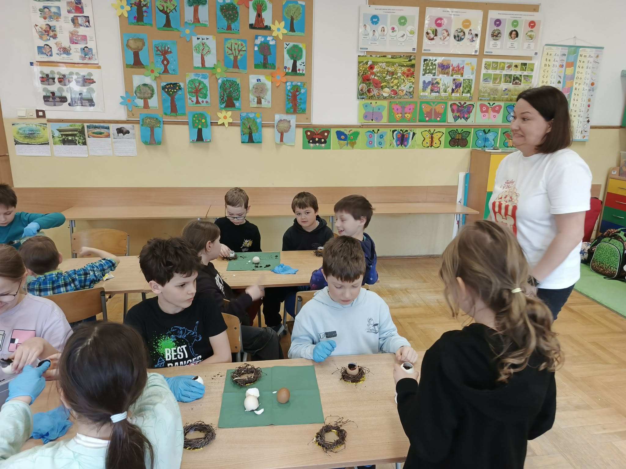 Dzieci tworzą świąteczne stroiki z wydmuszek i rzeżuchy. Na stołach leżą małe gniazdka z gałązek, a przy nich dzieci dekorują jajka i układają rośliny w wydmuszkach. W tle widać kolorową klasę z plakatami i pracami plastycznymi.