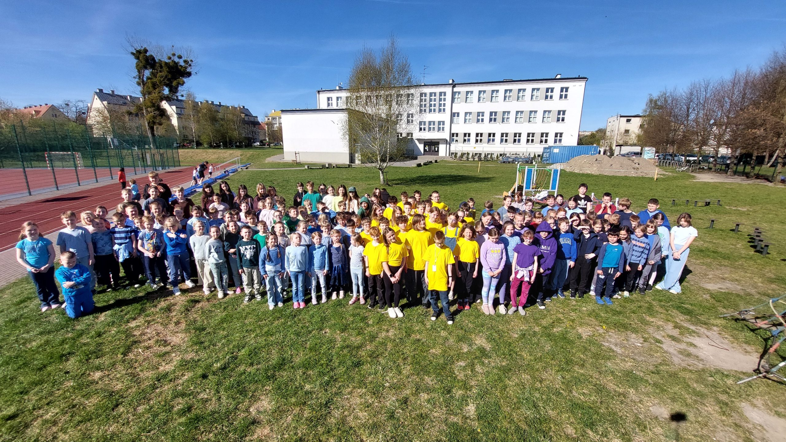 Szerokie ujęcie grupy uczniów stojących na trawniku przed białym, wielopiętrowym budynkiem szkoły. Dzieci są ubrane w kolorowe koszulki, z przewagą koloru żółtego i niebieskiego. Dzień jest słoneczny, niebo bezchmurne.