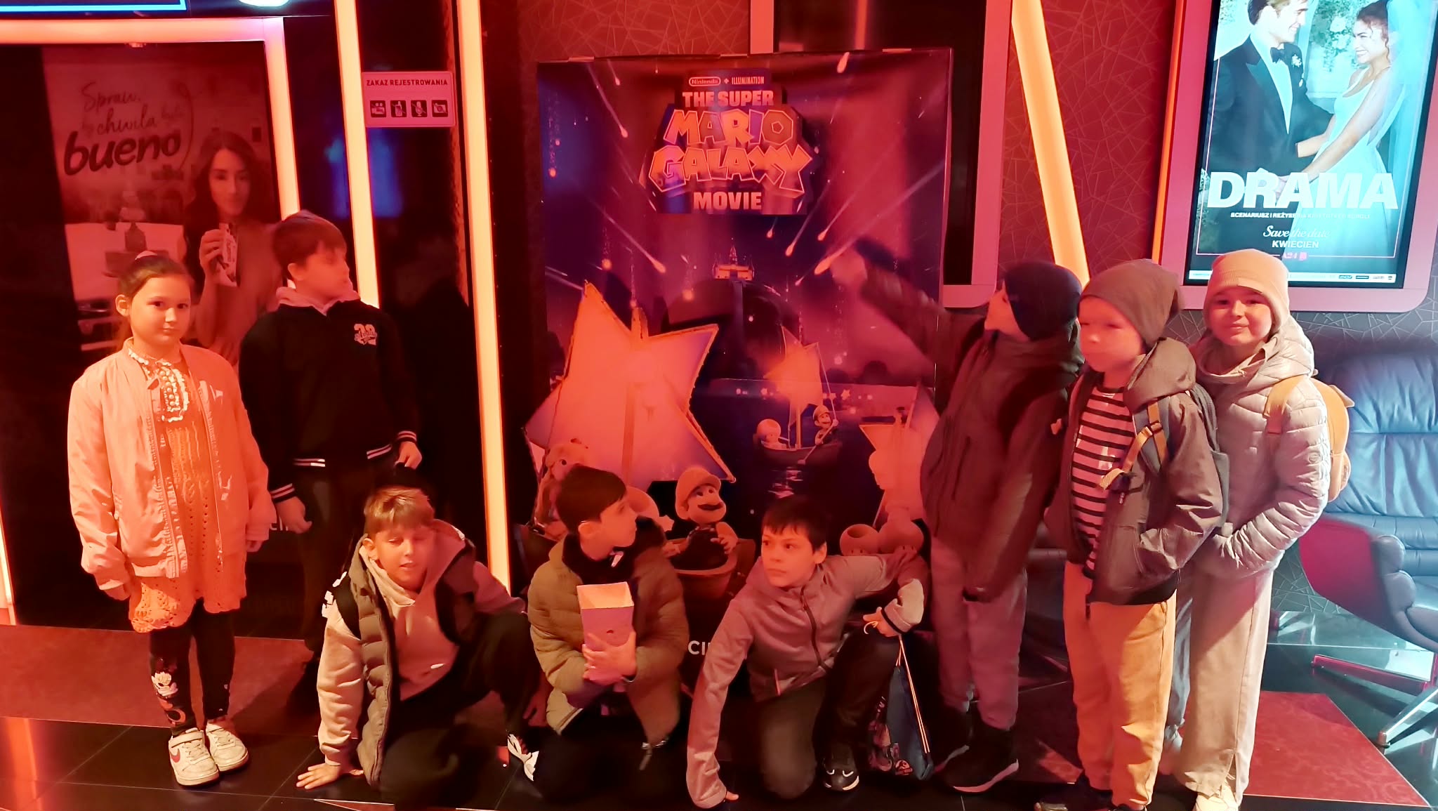 Uczniowie klasy 3c pozują w holu kina przy plakacie filmu animowanego „Super Mario Galaxy”; część dzieci trzyma popcorn, a wnętrze oświetlone jest czerwonym światłem.