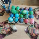 Karton z kolorowymi pisankami ustawionymi w rzędach. Pisanki są ozdobione wzorami wykonanymi woskiem pszczelim. Obok stoi kubek z kilkoma patyczkami do nakładania wosku, a pod pisankami leżą kartki z motywem jajek wielkanocnych.