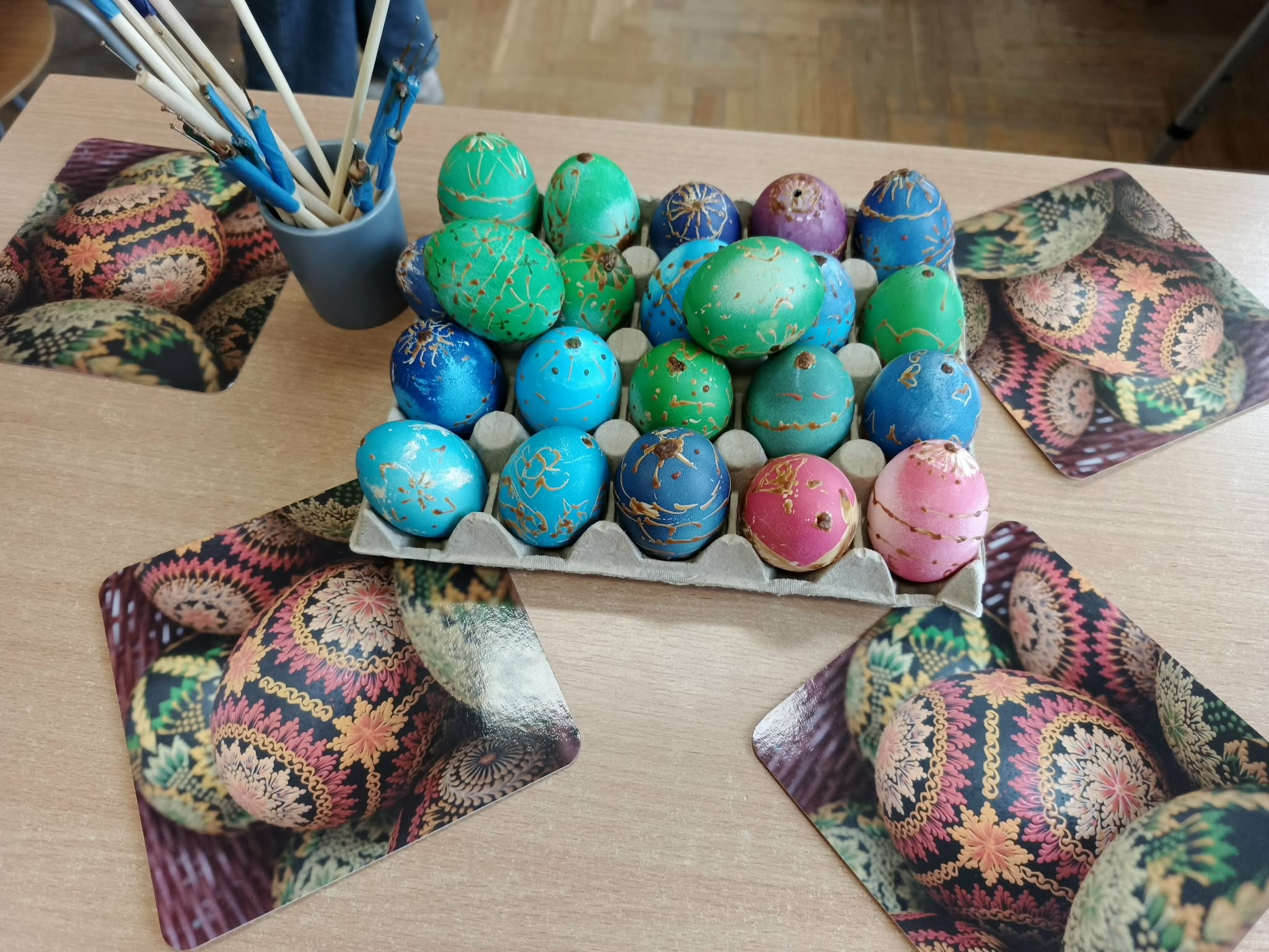 Karton z kolorowymi pisankami ustawionymi w rzędach. Pisanki są ozdobione wzorami wykonanymi woskiem pszczelim. Obok stoi kubek z kilkoma patyczkami do nakładania wosku, a pod pisankami leżą kartki z motywem jajek wielkanocnych.