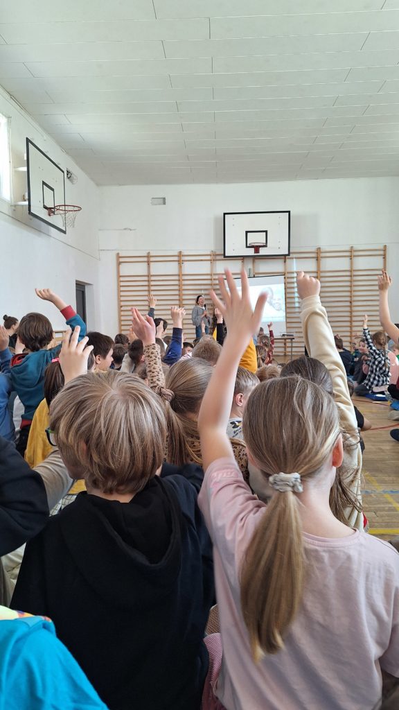 Warsztaty szkolne w sali gimnastycznej. Duża grupa uczniów siedzi na podłodze i aktywnie uczestniczy w spotkaniu – wielu z nich podnosi ręce, zgłaszając się do odpowiedzi. Z przodu stoi prowadząca przy ekranie z prezentacją.