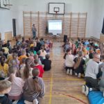 Sala gimnastyczna podczas warsztatów – uczniowie siedzą na materacach na podłodze, a prowadząca stoi przy ekranie i omawia prezentację dotyczącą Zespołu Downa.