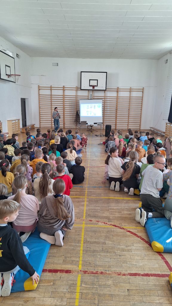 Sala gimnastyczna podczas warsztatów – uczniowie siedzą na materacach na podłodze, a prowadząca stoi przy ekranie i omawia prezentację dotyczącą Zespołu Downa.