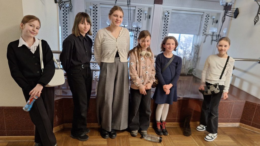 Sześć dziewczynek stoi obok siebie w eleganckim wnętrzu i pozuje do zdjęcia. Ubrane są w schludne ubrania. 