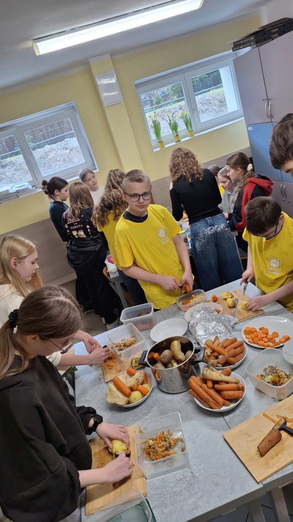 Grupa uczniów w szkolnej kuchni wspólnie przygotowuje posiłek, krojąc warzywa i pracując przy stołach pełnych składników.