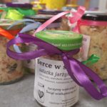 Słoiki z sałatką jarzynową ozdobione wstążkami stoją na stole, a w tle widać osobę w koszulce wolontariusza.