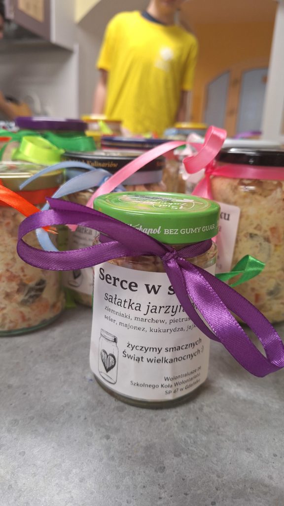 Słoiki z sałatką jarzynową ozdobione wstążkami stoją na stole, a w tle widać osobę w koszulce wolontariusza.