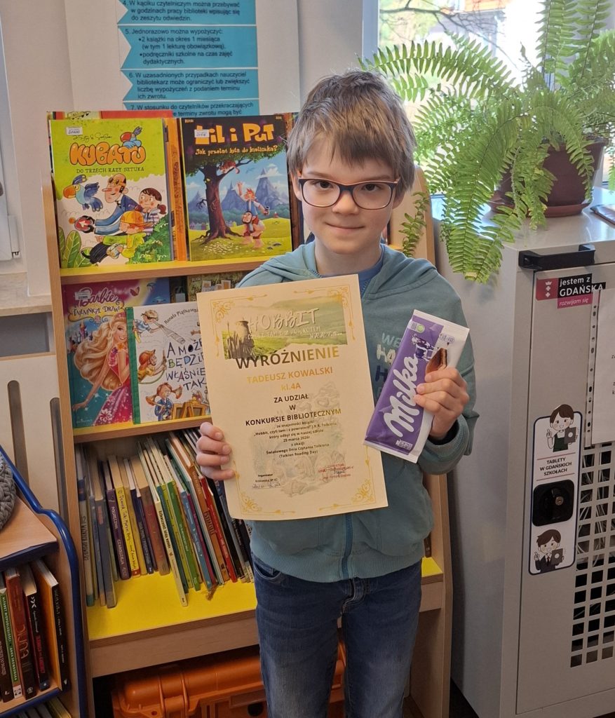 W centralnej części fotografii na tle regału bibliotecznego z książkami stoi uśmiechnięty chłopiec w okularach trzymający dyplom - wyróżnienie i czekoladę. 