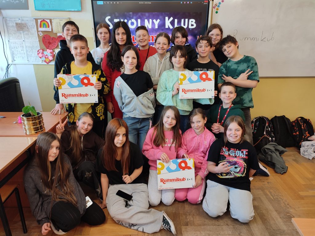 Grupa uczniów stoi przed tablicą interaktywną. Grupa jest uśmiechnięta, trzymają w rękach trzy opakowania gry Rummicub. W tle, na tablicy, jest napis Szkolny klub Rummikub.