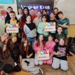 Grupa uczniów stoi przed tablicą interaktywną. Grupa jest uśmiechnięta, trzymają w rękach trzy opakowania gry Rummicub. W tle, na tablicy, jest napis Szkolny klub Rummikub.