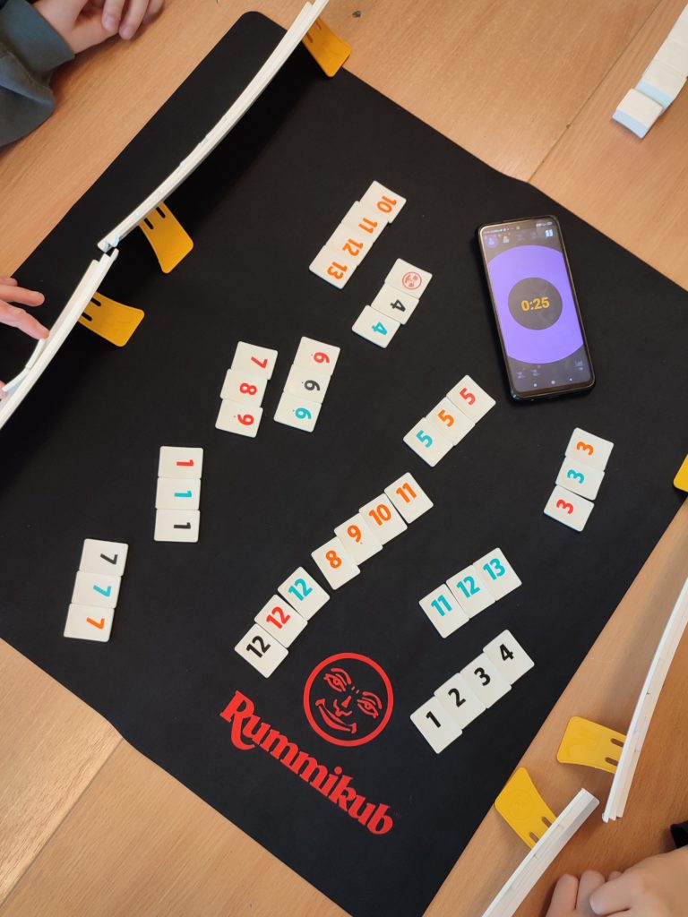 Widok z góry na złączone dwie ławki, na których leży mata Rummicub. Na macie rozgrywana jest gra: leży telefon odmierzający czas, leżą płytki z cyframi i liczbami oraz widoczne są cztery półeczki z płytkami graczy.