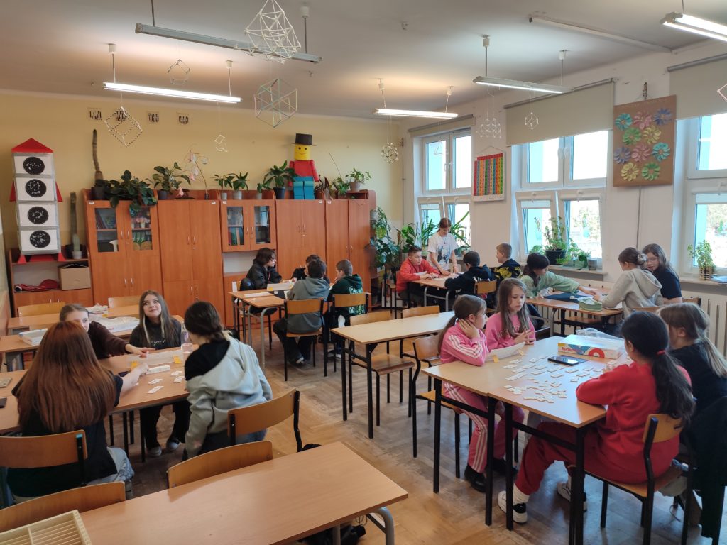 Szeroki widok klasy – W jasnej klasie kilka grup uczniów siedzi przy ławkach i gra w Rummikub. W tle widać szafki, rośliny oraz dekoracje geometryczne zawieszone pod sufitem.