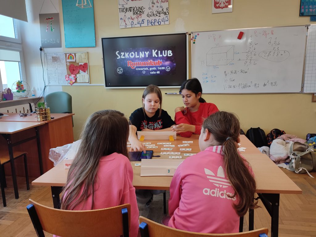 Gra przy tablicy interaktywnej – Cztery dziewczynki siedzą przy stole i grają w Rummikub, a za nimi znajduje się tablica z napisem „Szkolny Klub Rummikub”. Na ławce leżą płytki, a uczennice wspólnie analizują ruchy.