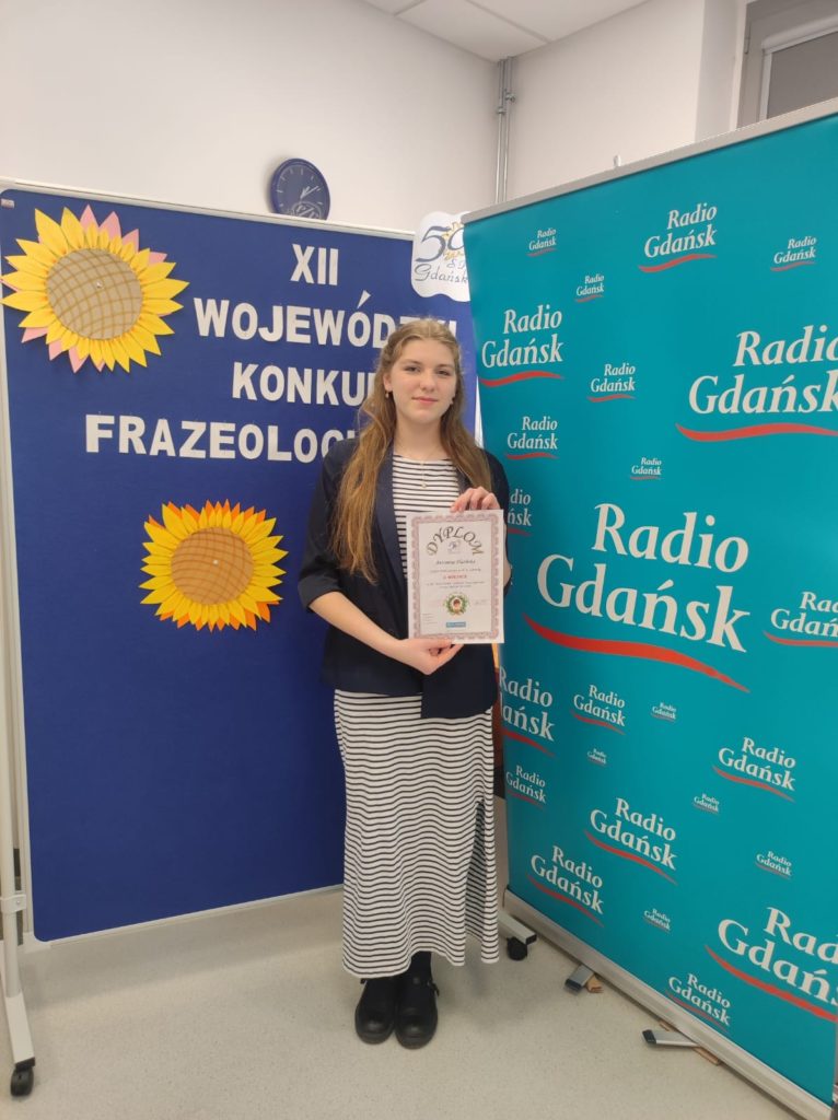 Tosia Flasińska stoi na tle dekoracji konkursowych i prezentuje swój dyplom.