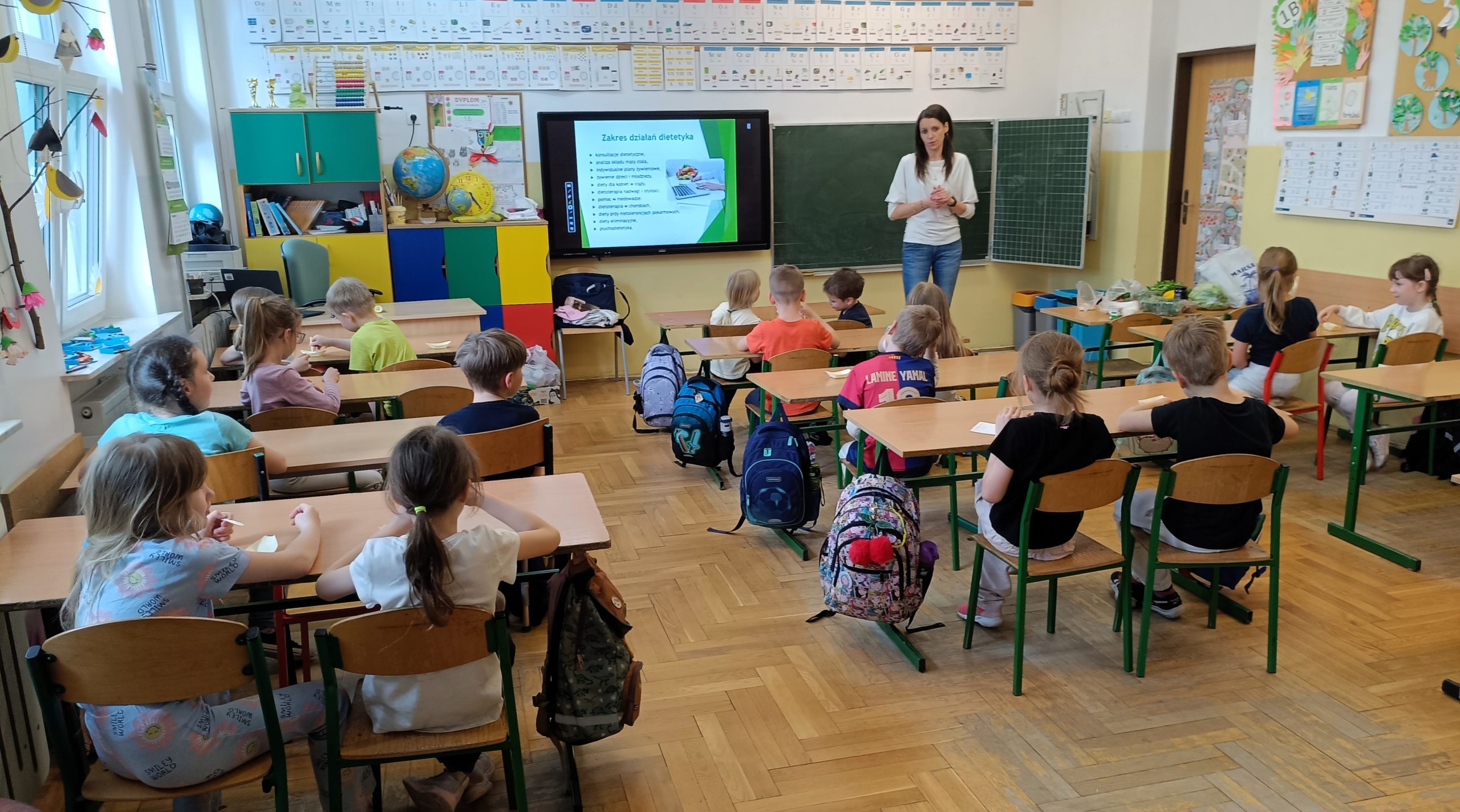 Pani stoi przy tablicy multimedialnej i prowadzi prezentację o zdrowym odżywianiu. Uczniowie siedzą w ławkach i słuchają zajęć.