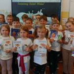 Na tle tablicy z napisem Światowy Dzień Mleka stoi ustawiona w trzy rzędy klasa Ic. W rękach uczniowie trzymają kocie zeszyty o mleku i kartonik mleka.