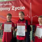 W sali gimnastycznej na tle plakatu Ogólnopolskiego Konkursu „Ratujemy Życie” stoją dwie uczennice i jeden uczeń SP 47 ubrani w czerwone koszulki reprezentacyjne. Z zadowoloną miną trzymają w dłoniach dyplomy.