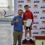 Mężczyzna i chłopiec pozują na tle białej ścianki z logo Gdańskiej Szkoły Szermierki (GSS) oraz herbem Gdańska. Chłopiec w czerwonej bluzie sportowej i czarnych spodenkach stoi na podium z numerem 2, trzymając w rękach złoty puchar oraz dyplom. Obok podium stoi uśmiechnięty mężczyzna w niebieskiej bluzie i jasnych spodniach, który pokazuje kciuk w górę. W tle widać fragment hali sportowej.