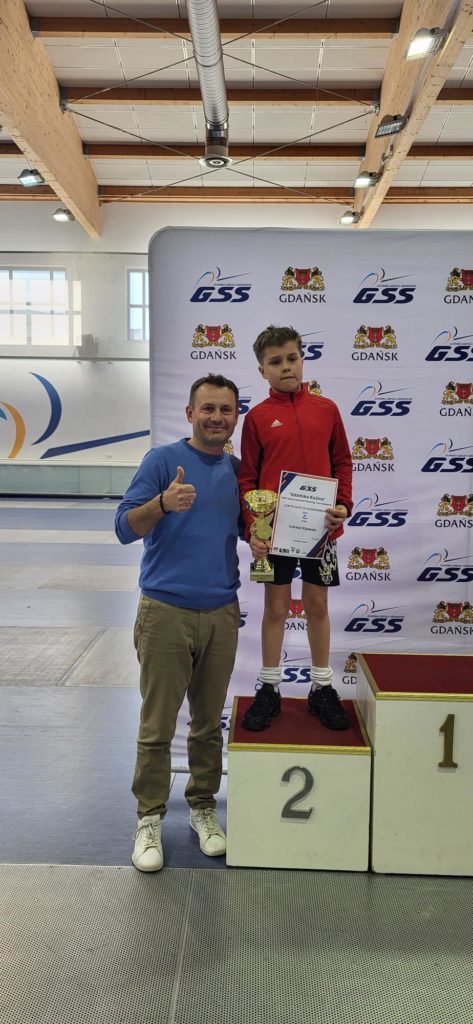 Mężczyzna i chłopiec pozują na tle białej ścianki z logo Gdańskiej Szkoły Szermierki (GSS) oraz herbem Gdańska. Chłopiec w czerwonej bluzie sportowej i czarnych spodenkach stoi na podium z numerem 2, trzymając w rękach złoty puchar oraz dyplom. Obok podium stoi uśmiechnięty mężczyzna w niebieskiej bluzie i jasnych spodniach, który pokazuje kciuk w górę. W tle widać fragment hali sportowej.