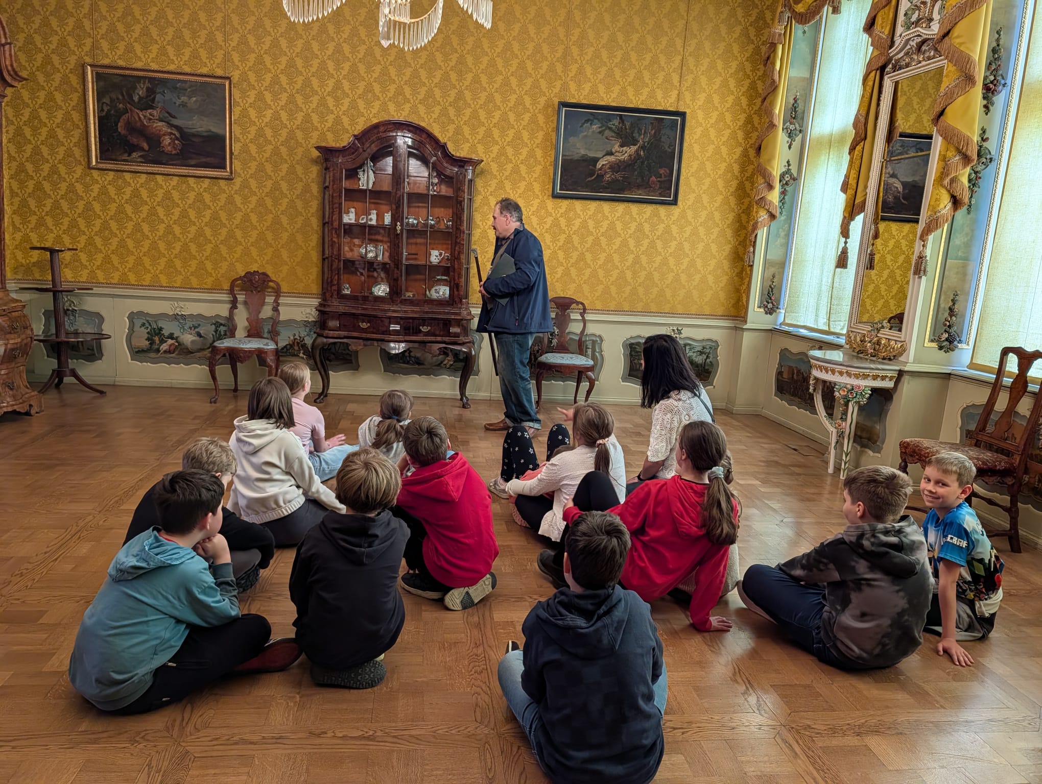  W sali muzealnej są dzieci , nauczycielka i pan który opowiada. Nauczycielka i dzieci siedzą na podłodze słuchają co mówi Pan prowadzący warsztaty edukacyjne. Po prawej stronie widać dwa duże okna. Na dużej ścianie widać dwa obrazy i komodę. Po prawej i lewej stronie komody są dwa krzesła.