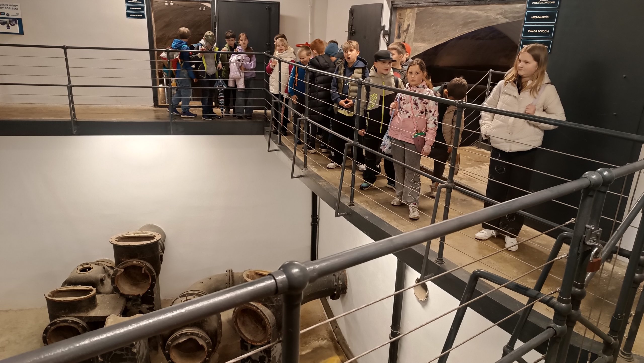 Widok z góry na metalową galeryjkę (pomost) z barierki, na której stoi grupa dzieci. Patrzą oni w dół, na dno betonowego wgłębienia, gdzie wyeksponowano stare, grube, czarne rury i zawory wodociągowe.