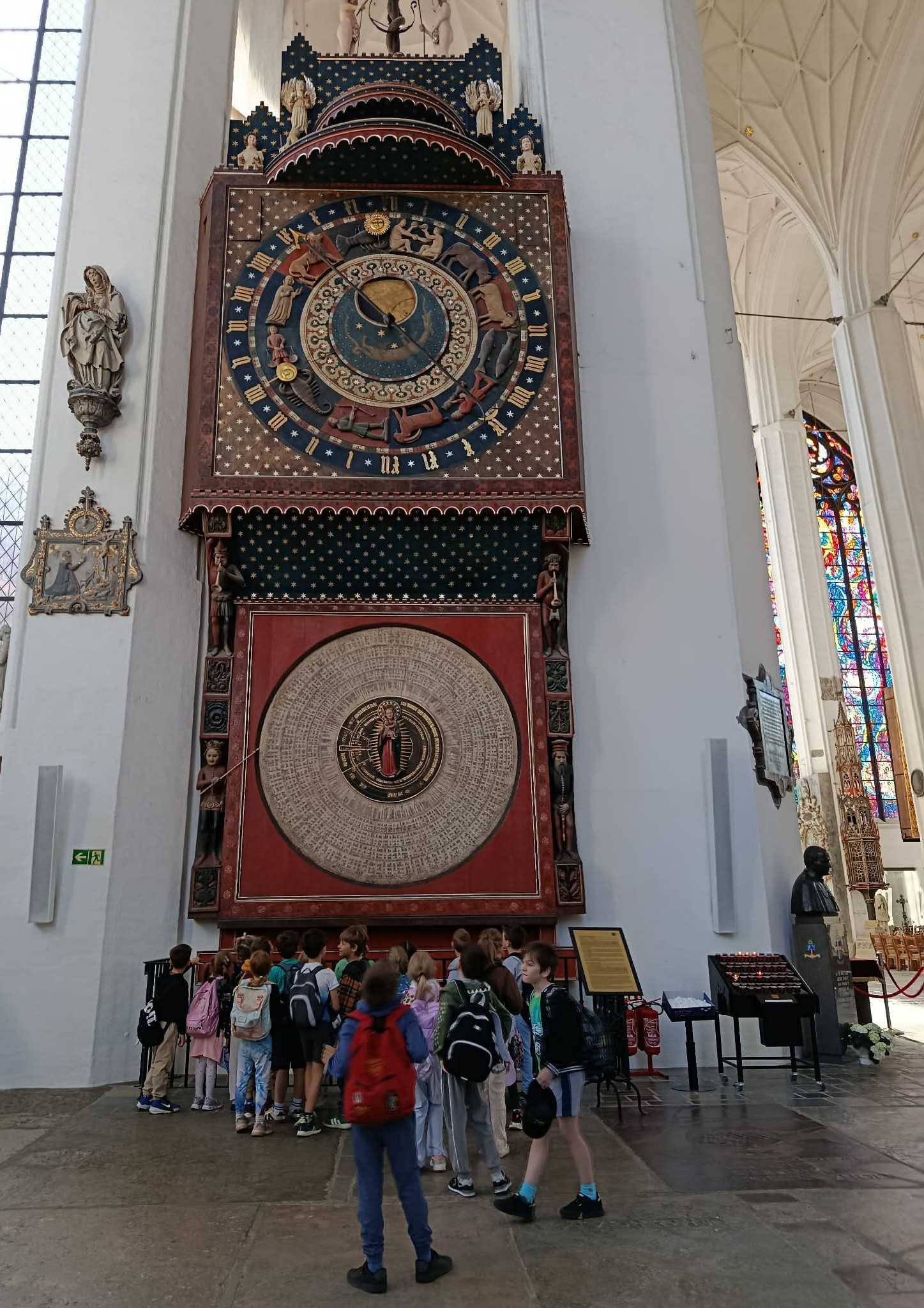 Na tle zegara astronomicznego w Bazylice Mariackiej stoi ustawiona tyłem grupa uczniów klasy 1c.