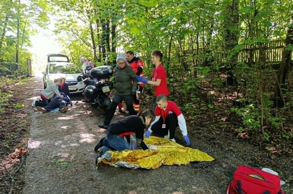Na leśnej drodze pozoracja wypadku z udziałem samochodu i motocykla. Dwie uczennice zajmują się motocyklistą, jeden uczeń uspokaja świadka i dwóch uczniów udziela pomocy niemowlęciu. Przy nich ratownik oceniający ich działania.