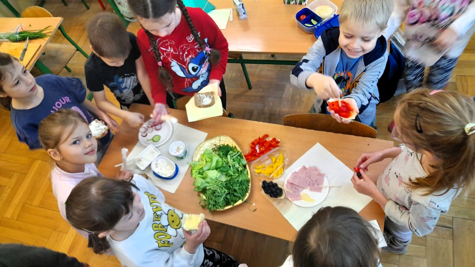 Grupa dzieci przygotowuje zdrowe kanapki przy wspólnym stole. Na stole znajdują się warzywa, pieczywo i dodatki.