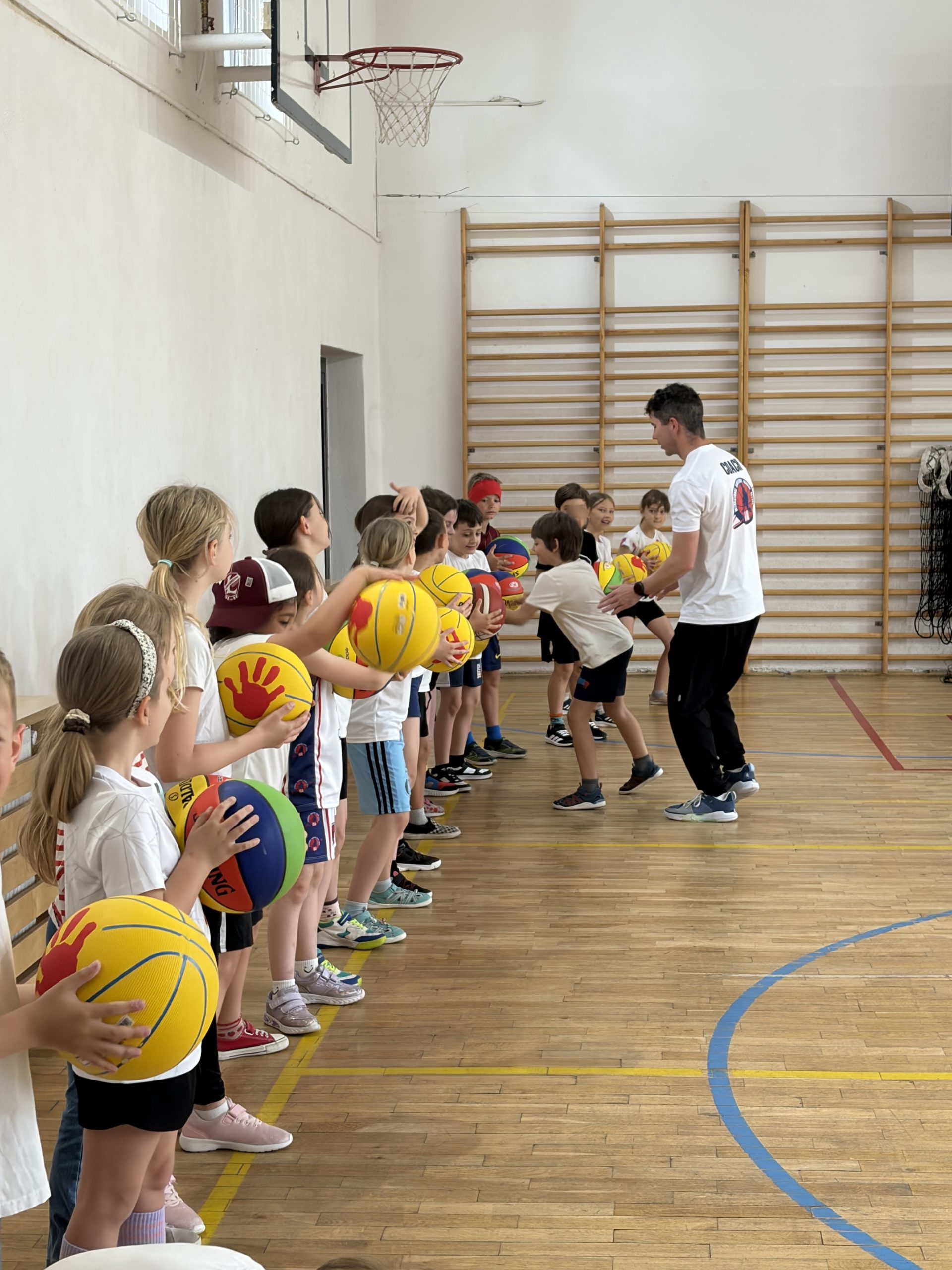 Kolejka uczniów stojąca przy drabinkach gimnastycznych, każdy trzyma w rękach kolorową piłkę do koszykówki. Trener w koszulce "COACH" rozdaje lub instruuje jednego z uczniów, a inne dzieci patrzą na nich.