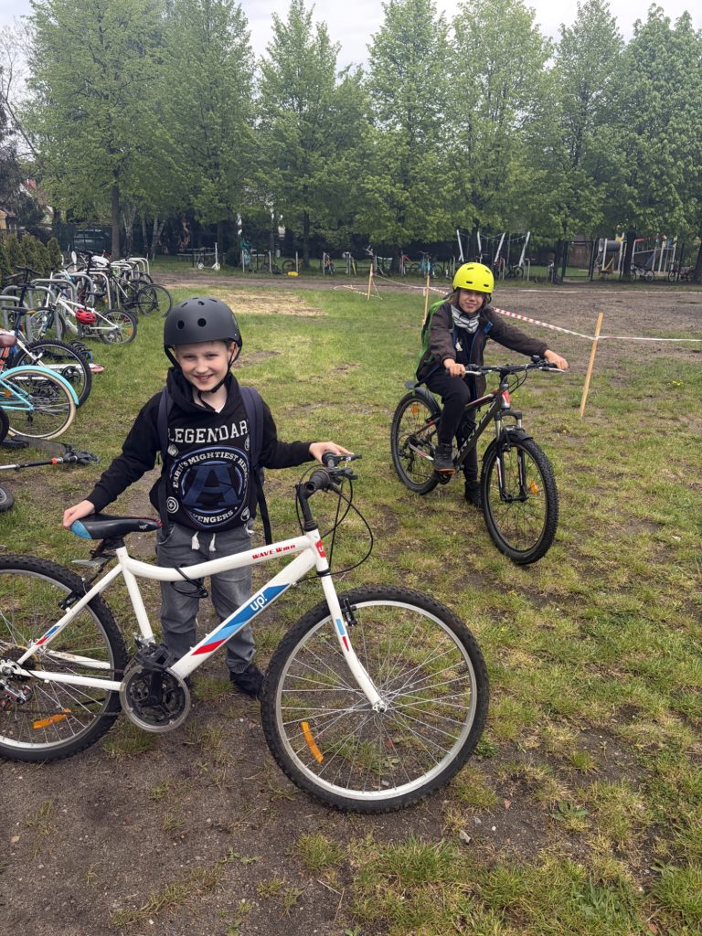 Dwaj chłopcy stoją na trawiasto-piaszczystym parkingu rowerowym w kaskach i uśmiechają się do obiektywu. Jeden trzyma rękę na białym rowerze, drugi siedzi na czarnym rowerze górskim. W tle widać rzędy rowerów i dużo zielonych drzew.