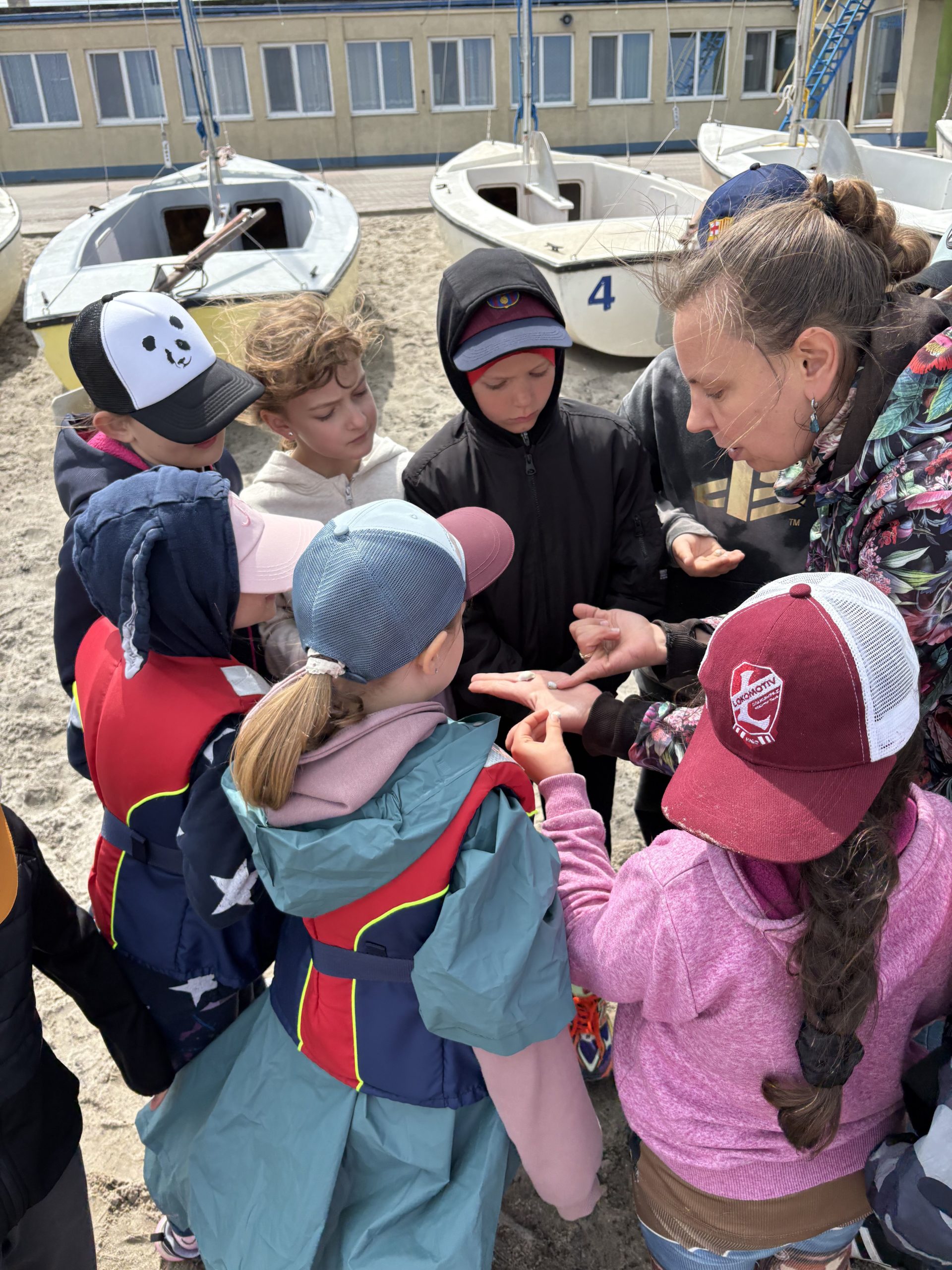 Kobieta pokazuje grupie dzieci zebranych na plaży drobne znaleziska trzymane na otwartej dłoni, w tle stoją białe łódki.