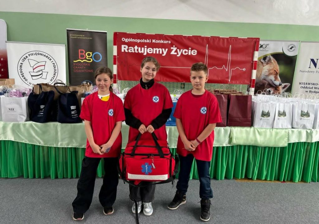W sali gimnastycznej na tle plakatu Ogólnopolskiego Konkursu „Ratujemy Życie” i nagród ustawionych na zielonym stole stoją dwie uczennice i jeden uczeń SP 47 ubrani w czerwone koszulki reprezentacyjne. Jedna z uczennic trzyma apteczkę.