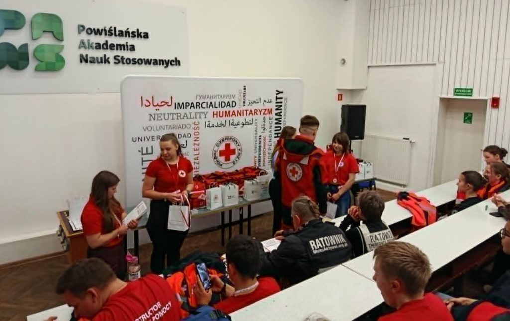 Na tle baneru PCK uczennica SP 47 w Gdańsku odbiera medal i nagrodę najlepszego ratownika XXXII Ogólnopolskich Mistrzostw Pierwszej Pomocy PCK. Obok organizatorzy mistrzostw, nagrody oraz siedzący na auli uczestnicy.