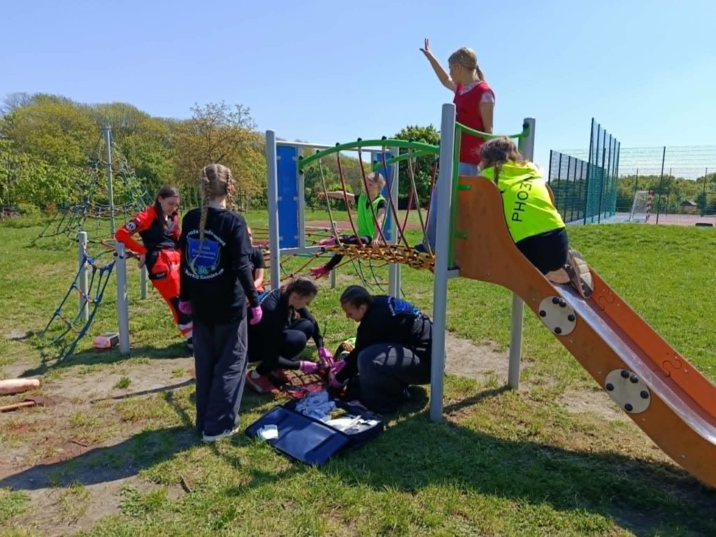 Na szkolnym placu zabaw na tle zjeżdżalni trzyosobowa drużyna dziewcząt opatrująca amputację nogi podczas pozoracji zdarzenia. Przy nich świadkowie stojący na zjeżdżalni i dwie ratowniczki oceniające udzielanie pomocy.